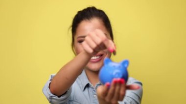 Mavi kot gömlekli mutlu kadın domuz kumbarasına bozuk para koyuyor, baş parmak işareti yapıyor ve sarı arka planda gülümsüyor.