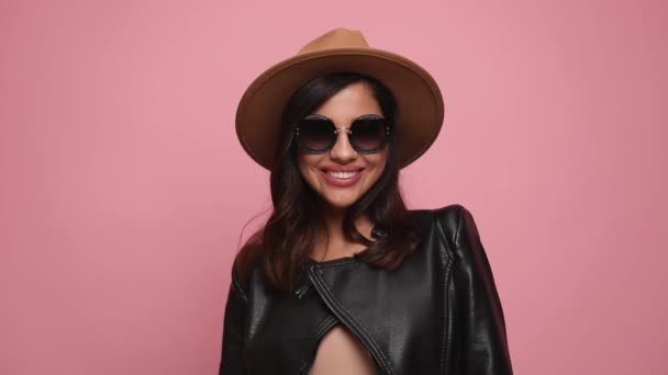 穿着皮夹克 头戴帽子 戴着太阳镜 面带微笑 头戴眼镜 头戴粉色背景的时尚女人 — 图库视频影像