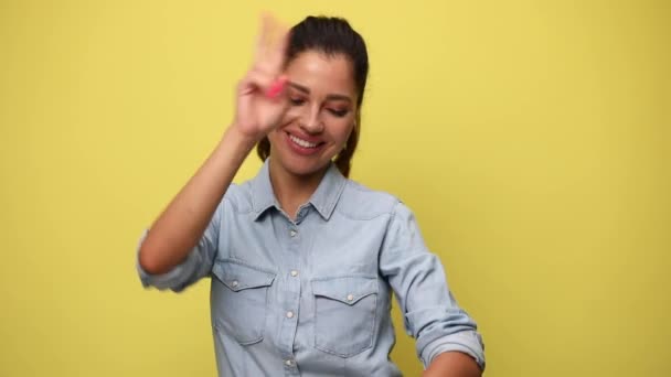 青いジーンズのシャツを着た幸せな女の子がダンスのジェスチャーを作り 空気中で手を取り 黄色の背景で踊り — ストック動画