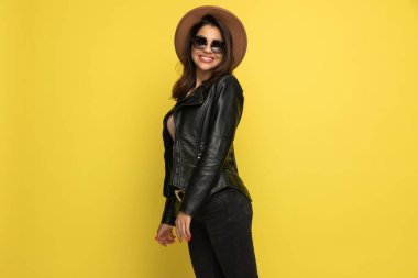 Deri ceketli, güneş gözlüklü, şapkalı, gülümseyen, sarı arka planda yan görünüm pozisyonunda duran seksi bir kadın.