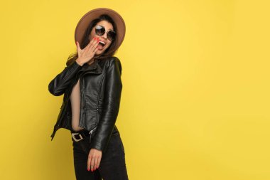 Deri ceketli, ağzı eliyle kaplayan ve sarı arka planda poz veren havalı bir kız.
