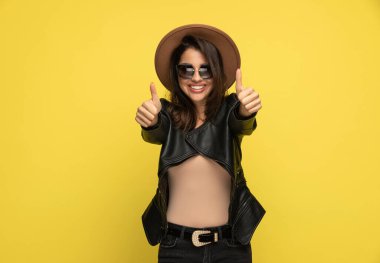Deri ceketli, güneş gözlüklü ve şapkalı mutlu bir kadın. Baş parmak hareketi yapıyor ve sarı arka planda gülümsüyor.