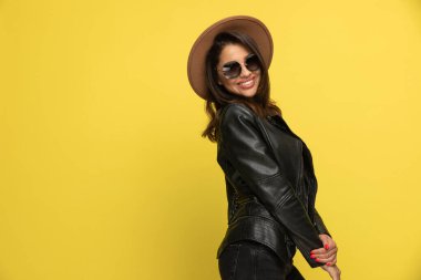 Deri ceketli, güneş gözlüğü ve şapka takan, el ele tutuşan ve gülümseyen, sarı arka planda duran seksi genç kadın.