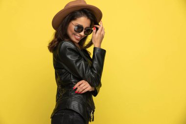 Deri ceketli, şapka ve güneş gözlüğü takan, saçları düzenleyen ve sarı arka plana bakan ürkek genç bir kız.