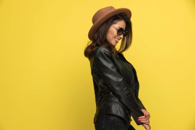 Deri ceketli utangaç genç kız aşağı bakıyor ve sarı arka planda el ele tutuşuyor.