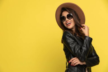 Deri ceketli seksi genç bir kadın saçını tutuyor ve sarı arka planda gülümsüyor.