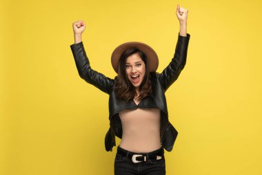 Deri ceketli neşeli kadın kafasının üstünde yumruklar tutuyor ve sarı arka planda kutlama yapıyor.