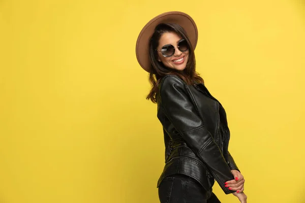 穿着皮夹克 戴着太阳镜和帽子 手牵手 微笑着 站在一个黄色背景的侧面的性感年轻女人 — 图库照片