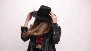 Havalı kız hareket ediyor, dans ediyor ve eğleniyor, aşağı bakıyor ve şapkasını düzeltiyor, gri arka planda gülüyor.