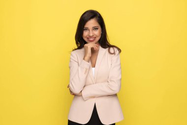 Pembe elbiseli genç iş kadını kollarını kavuşturuyor, el ele tutuşuyor ve düşünüyor, gülümsüyor ve sarı arka planda duruyor.