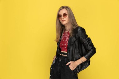 İki elini de cebinde tutan, sarı stüdyo arka planında dururken güneş gözlüğü takan kendine güvenen bir manken.