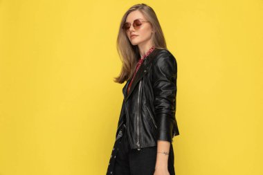 Stüdyonun sarı arka planında dururken gülümseyen ve güneş gözlüğü takan manken.