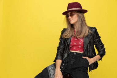 Şapka ve güneş gözlüğü takan havalı bir manken, sarı stüdyo arka planında bir sandalyede otururken el ele tutuşuyor.