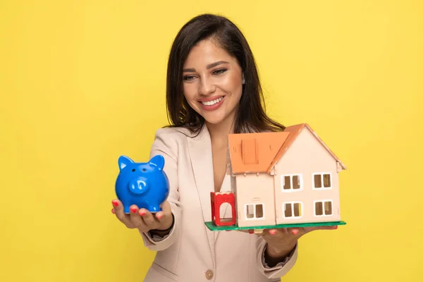 young girl holding and recommending piggy bank and building model, smiling on yellow background