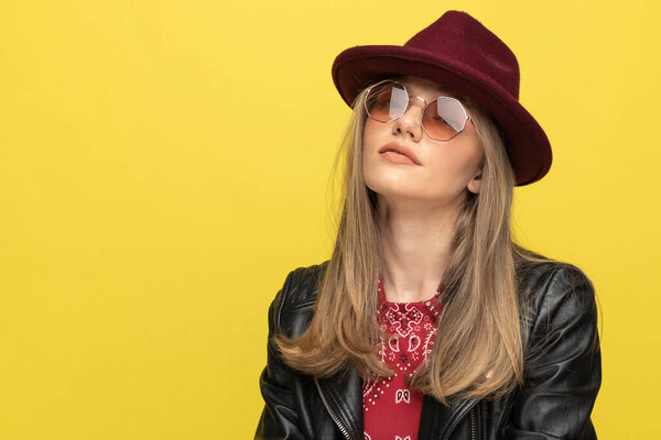 Closeup of serious fashion model wearing hat and sunglasses while standing on yellow studio background