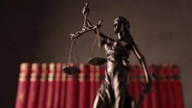 Gözleri bağlı bir kadın tarafından temsil edilen adalet heykeli ve gücü ve tarafsızlığı simgeleyen bir kılıç kahverengi arka planda kırmızı kitapların önünde dönüyor.