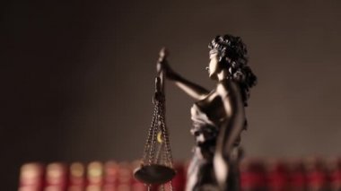 Gözleri bağlı bir kadın tarafından temsil edilen adalet heykeli ve gücü ve tarafsızlığı simgeleyen bir kılıç kahverengi arka planda kırmızı kitapların önünde dönüyor.