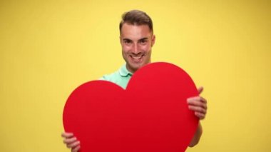 Büyük kırmızı kalbini gösteren, kucaklayan ve sarı arka planı onaylayan sıradan bir adam.