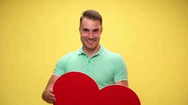 Genç adam yavaşça büyük kırmızı kalbini gösteriyor ve sarı arka plandaki kameraya veriyor.