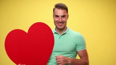 Elinde kocaman kırmızı bir kalp tutan seksi bir adam, kamerayı işaret ediyor, bana işaret ediyor ve sarı arka plandaki kalbi işaret ediyor.