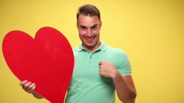 Elinde kocaman kırmızı bir kalp tutan çekici bir adam, kameraya ve kendisine bakıyor ve sarı arka planda sen ve ben işareti yapıyor.