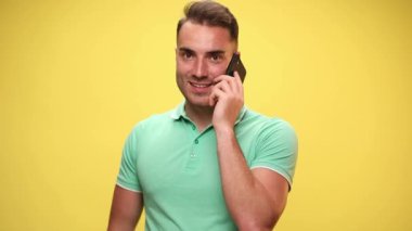 Yakışıklı sıradan bir adam cep telefonuyla konuşuyor, etrafa bakıyor, gülümsüyor ve sarı arka planda eğleniyor.