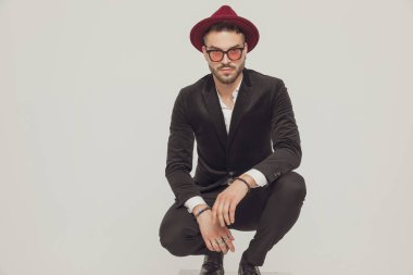 Gri stüdyo arka planında çömelirken şapka ve güneş gözlüğü takan kendine güvenen bir model.