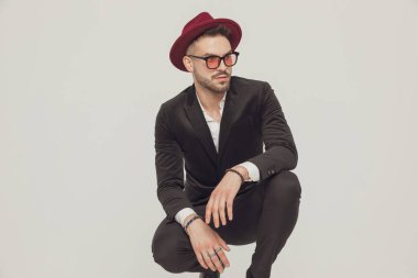 Moda mankeni gözlerini kaçırıyor, şapka ve güneş gözlüğü takıyor ve gri stüdyo arka planında çömeliyor.