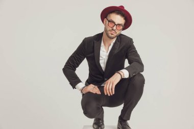 Üzgün model ileriye bakıyor, güneş gözlüğü ve şapka takıyor ve gri stüdyo arka planında çömeliyor.