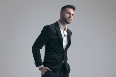 Gri stüdyo arka planında yürürken iki eli ceplerinde, takım elbiseli, canı sıkkın, zarif bir adam.