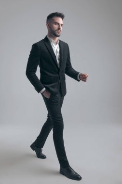 Gri stüdyo arka planında yürürken takım elbise giyen, el ele tutuşan kararlı, zarif bir adam.