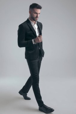 Takım elbise giyen ve gri stüdyo geçmişinde yürüyen sert, zarif bir adam.