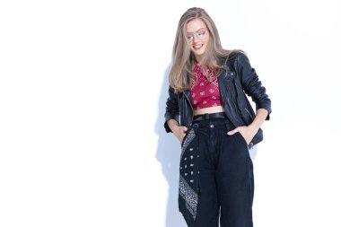 Neşeli bir manken, iki eli ceplerinde, gözlüklü ve deri ceketli beyaz stüdyo arka planında dururken gülüyor.