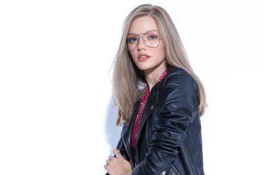 Çekici bir manken, beyaz stüdyo arka planında dikilirken kollarını düzeltiyor, gözlük ve deri ceket giyiyor.