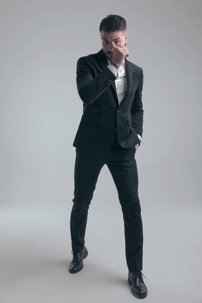 Handsome elegant man holding hand in pocket and scratching nose, wearing suit while walking on gray studio background