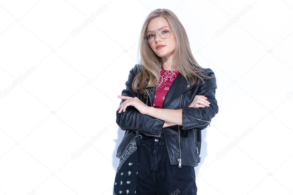 Charming fashion model holding hand crossed, wearing glasses and leather jacket while standing on white studio background