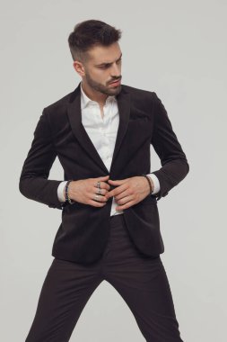 Gri stüdyo arka planında bacaklarını açarak dikilirken ceketinin düğmelerini çözüyor.