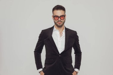 Gri stüdyo arka planında dururken iki elini de cebinde tutan ve güneş gözlüğü takan kendine güvenen bir manken.