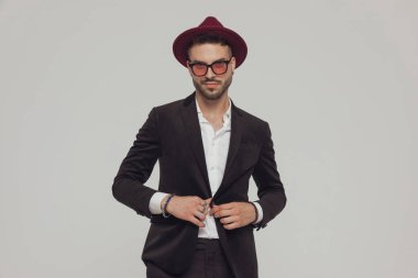 Pozitif model model ceketi, şapka ve güneş gözlüğü takıyor, gülümsüyor ve gri stüdyo arka planında duruyor.