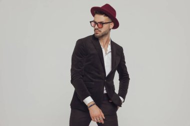 Kendine güveni tam bir manken. Gri stüdyo arka planında dururken el ele bakıyor, şapka ve güneş gözlüğü takıyor.