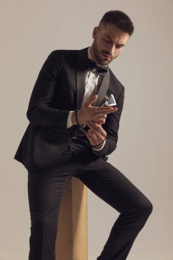 Düşünceli damat el ele tutuşuyor ve onlara bakıyor, gri stüdyo arka planında otururken smokin giyiyor.