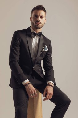 Şüpheli damat, gri stüdyo arka planında otururken smokin giyip ileriye bakıyor.