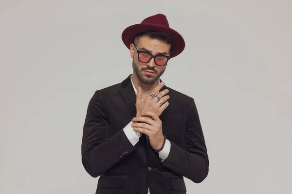 魅力的なファッションモデルは グレーのスタジオの背景に立っている間 帽子とサングラスを身に着けて 彼の袖を配置 — ストック写真