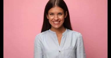 Mavi kot gömlek giyen, kollarını kavuşturup pembe arka planda gülümseyen genç bir kadın.