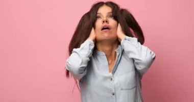 Uyanık kalmaya çalışan, pembe arka planda rahatsız edici bir şekilde çenesine dokunan, kot gömlekli, uykulu, sıradan bir kadın.