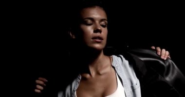 Sim kaplı deri ceketli seksi kız omuzlarından ceketini çıkartıyor, yan görüş pozisyonunda poz veriyor, el hareketleri yapıyor, siyah arka planda ceket ayarlıyor.