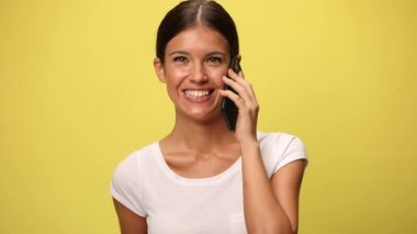 Genç, gülümseyen, sıradan bir kadın cep telefonuyla konuşuyor ve sarı arka planı onaylıyor.