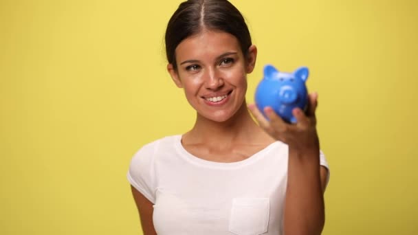 若いカジュアルな女性が貯金箱を持って黄色の背景にお金を入れ — ストック動画