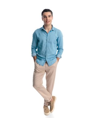 Mavi kotlu gömlekli yakışıklı genç adam ceplerinde el ele tutuşuyor, beyaz arka planda duruyor ve ileriye bakıyor.