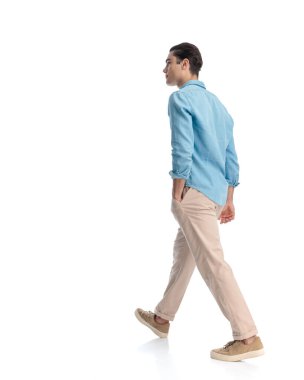 Mavi kot gömlekli seksi genç adam beyaz arka plana bakarken elini cebine atarak ilerliyor.